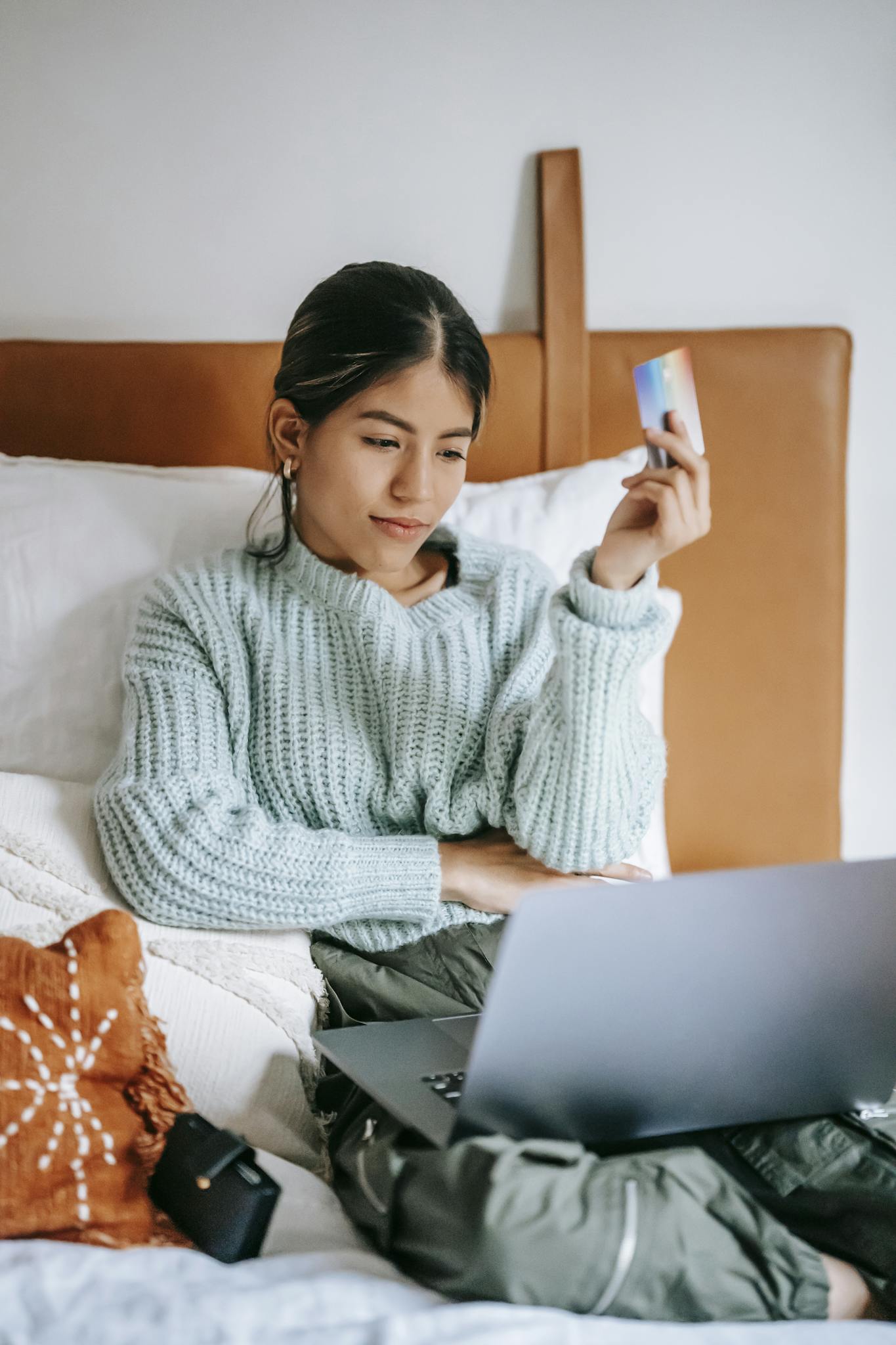 A young woman relaxes in bed, shopping online with a laptop and credit card in hand.